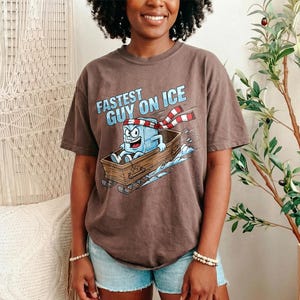 May include: A brown t-shirt featuring the phrase "Fastest Guy on Ice" and a cartoon ice cube character on a sled. The ice cube is wearing a red and white scarf. The shirt is worn with blue denim shorts.