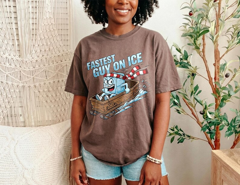 May include: A brown t-shirt featuring the phrase "Fastest Guy on Ice" and a cartoon ice cube character on a sled. The ice cube is wearing a red and white scarf. The shirt is worn with blue denim shorts.