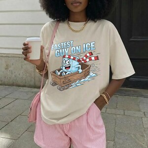 May include: Beige t-shirt with the text "Fastest Guy on Ice" and a cartoon ice cube character in a wooden sled. The ice cube wears a red and white scarf. The person is wearing pink and white striped shorts.