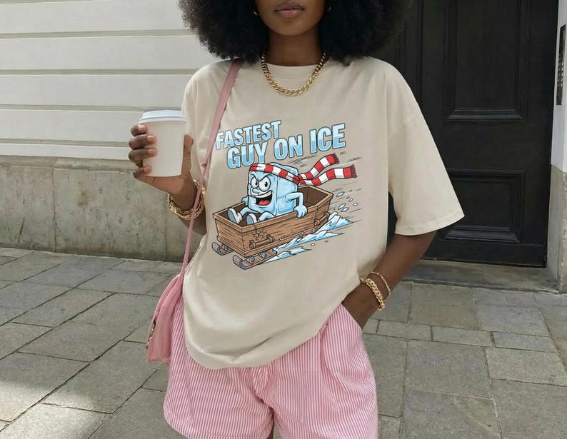 May include: Beige t-shirt with the text "Fastest Guy on Ice" and a cartoon ice cube character in a wooden sled. The ice cube wears a red and white scarf. The person is wearing pink and white striped shorts.