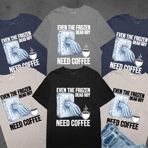 May include: A collection of t-shirts in various colors, including navy, gray, and black. Each shirt features the text "EVEN THE FROZEN DEAD GUY NEED COFFEE" with a graphic of a hand reaching out of ice to hold a coffee cup.