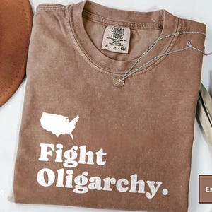 May include: A brown t-shirt with a white outline of the United States and the text "Fight Oligarchy." The shirt is folded and laid flat on a white surface. The word "Espresso" is visible in the bottom left corner of the image.