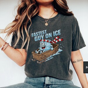 May include: Dark grey t-shirt featuring the text "Fastest Guy on Ice" and a cartoon ice cube character on a wooden sled. The ice cube wears a red and white striped scarf and has a determined expression.