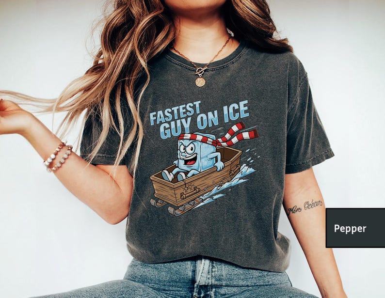 May include: Dark grey t-shirt featuring the text "Fastest Guy on Ice" and a cartoon ice cube character on a wooden sled. The ice cube wears a red and white striped scarf and has a determined expression.