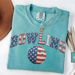 May include: A seafoam green short-sleeve t-shirt featuring the word BOWLING in a red, white, and blue American flag pattern. Below the text is a circular bowling ball graphic with the same flag design. The shirt has a Comfort Colors brand label.