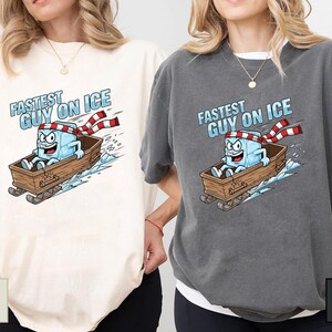May include: Ivory and pepper-coloured t-shirts with a cartoon ice cube character in a wooden sled, text reads "Fastest Guy on Ice." The ice cube wears a red and white striped scarf and has a determined expression.