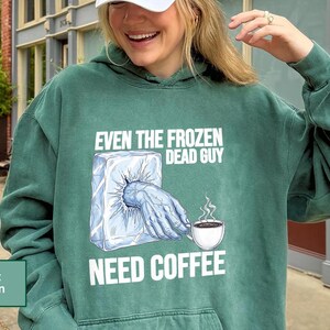 May include: Light green hoodie with the text "EVEN THE FROZEN DEAD GUY NEED COFFEE" and a graphic of a hand reaching out of an ice block to hold a coffee cup. The hoodie has a drawstring hood and a kangaroo pocket.