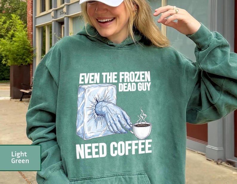 May include: Light green hoodie with the text "EVEN THE FROZEN DEAD GUY NEED COFFEE" and a graphic of a hand reaching out of an ice block to hold a coffee cup. The hoodie has a drawstring hood and a kangaroo pocket.