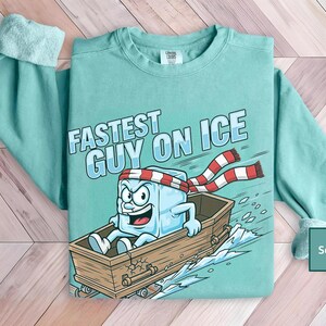 May include: Seafoam green sweatshirt featuring the text "Fastest Guy on Ice" and a cartoon ice cube character in a wooden boat. The ice cube wears a red and white striped scarf and headband. The sweatshirt has long sleeves.