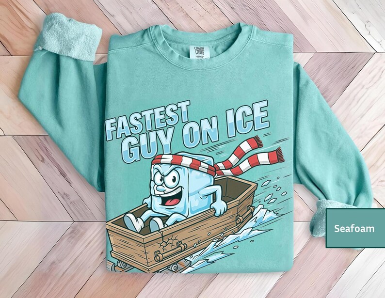 May include: Seafoam green sweatshirt featuring the text "Fastest Guy on Ice" and a cartoon ice cube character in a wooden boat. The ice cube wears a red and white striped scarf and headband. The sweatshirt has long sleeves.