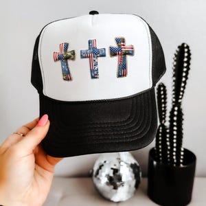 May include: A black and white trucker hat featuring three embroidered cross patches on the front panel. Each cross is decorated with a patchwork design incorporating American flag patterns, camouflage, and denim-like textures in blue, red, and white.
