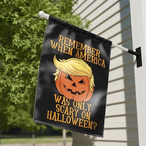 Puede incluir: Bandera negra de Halloween con el texto "REMEMBER WHEN AMERICA WAS ONLY SCARY ON HALLOWEEN?" y una calabaza con pelo amarillo. La bandera est&aacute; colgada afuera en un poste blanco.