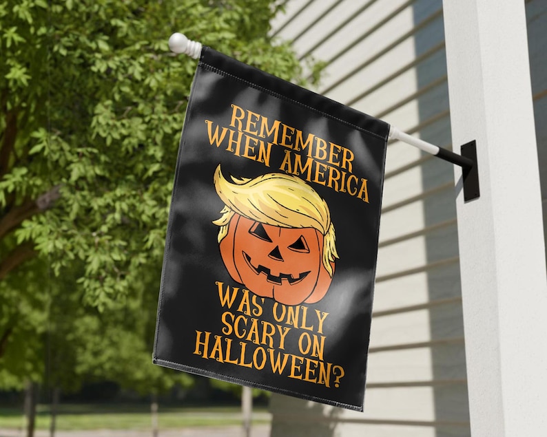Puede incluir: Bandera negra de Halloween con el texto "REMEMBER WHEN AMERICA WAS ONLY SCARY ON HALLOWEEN?" y una calabaza con pelo amarillo. La bandera est&aacute; colgada afuera en un poste blanco.