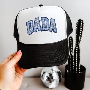 May include: A trucker hat with a white front panel and black mesh back. The front features the word DADA in blue and white gingham patterned letters with a distressed, stitched-style outline. The brim and the front panel are solid black.