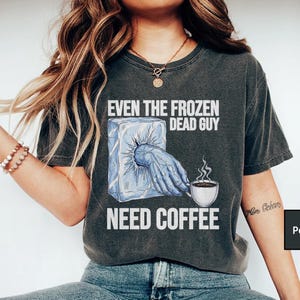 May include: Dark gray t-shirt with the text "EVEN THE FROZEN DEAD GUY NEED COFFEE." The graphic shows a hand reaching out of an ice cube to hold a coffee cup. The shirt is a comfortable, casual style.