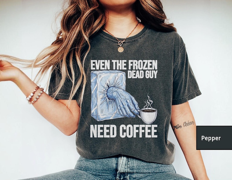 May include: Dark gray t-shirt with the text "EVEN THE FROZEN DEAD GUY NEED COFFEE." The graphic shows a hand reaching out of an ice cube to hold a coffee cup. The shirt is a comfortable, casual style.