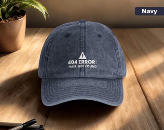 May include: Navy blue baseball cap with a curved brim. The cap features the text "404 ERROR: HAIR NOT FOUND" in white, with a white warning symbol. The hat is made of a textured fabric and sits on a wooden surface.