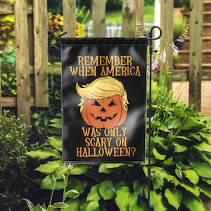 Puede incluir: Bandera de jard&iacute;n negra de Halloween con el texto "REMEMBER WHEN AMERICA WAS ONLY SCARY ON HALLOWEEN?" y una calabaza con pelo rubio. La bandera se muestra al aire libre frente a una valla de madera y follaje verde.