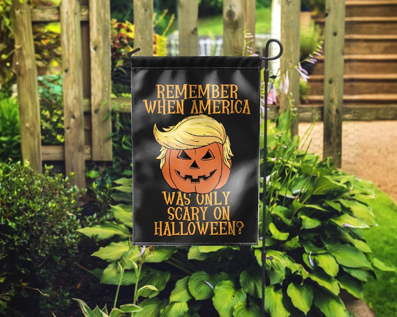 Puede incluir: Bandera de jard&iacute;n negra de Halloween con el texto "REMEMBER WHEN AMERICA WAS ONLY SCARY ON HALLOWEEN?" y una calabaza con pelo rubio. La bandera se muestra al aire libre frente a una valla de madera y follaje verde.