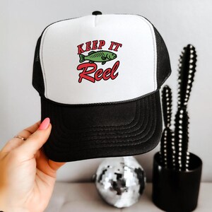 May include: Black and white trucker hat with the text "Keep It Reel" in red and green. The hat features a green fish graphic. The hat has a black brim and mesh back.
