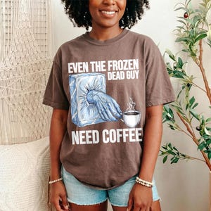 May include: A brown t-shirt with the text "EVEN THE FROZEN DEAD GUY NEED COFFEE". The graphic shows a frozen hand reaching out of a block of ice, with a coffee cup. The shirt is worn by a person.
