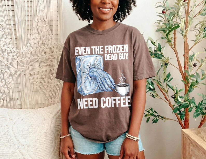 May include: A brown t-shirt with the text "EVEN THE FROZEN DEAD GUY NEED COFFEE". The graphic shows a frozen hand reaching out of a block of ice, with a coffee cup. The shirt is worn by a person.