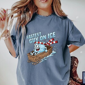 May include: Denim blue t-shirt featuring the text "Fastest Guy on Ice" and a cartoon ice cube character wearing a scarf and riding a sled. The ice cube has a surprised expression. The sled is brown.