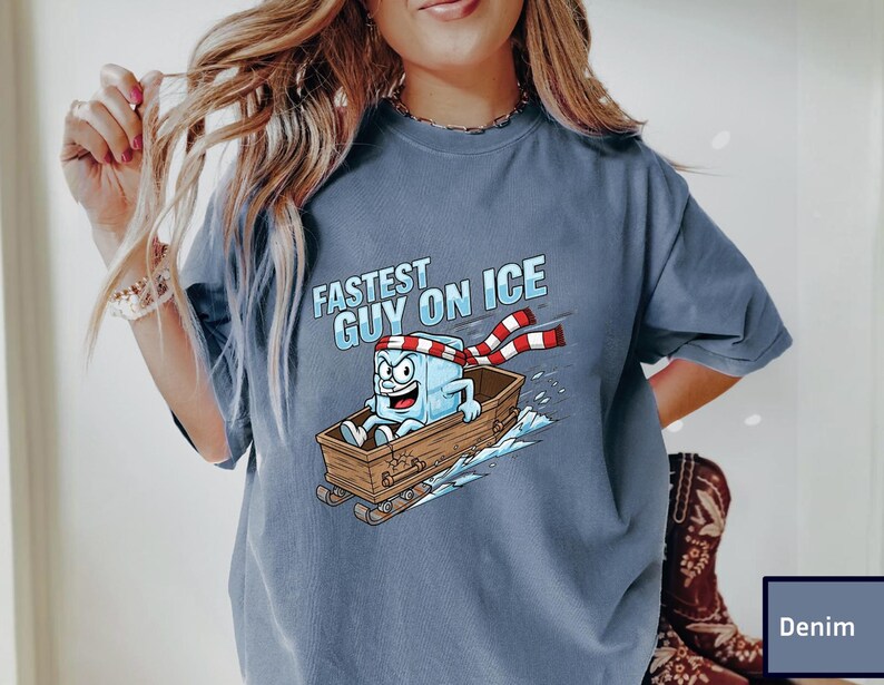 May include: Denim blue t-shirt featuring the text "Fastest Guy on Ice" and a cartoon ice cube character wearing a scarf and riding a sled. The ice cube has a surprised expression. The sled is brown.