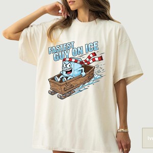 May include: Ivory t-shirt with a cartoon ice cube character in a wooden sled, with the text "FASTEST GUY ON ICE." The ice cube wears a red and white striped scarf and headband. The design includes blue ice and snow.