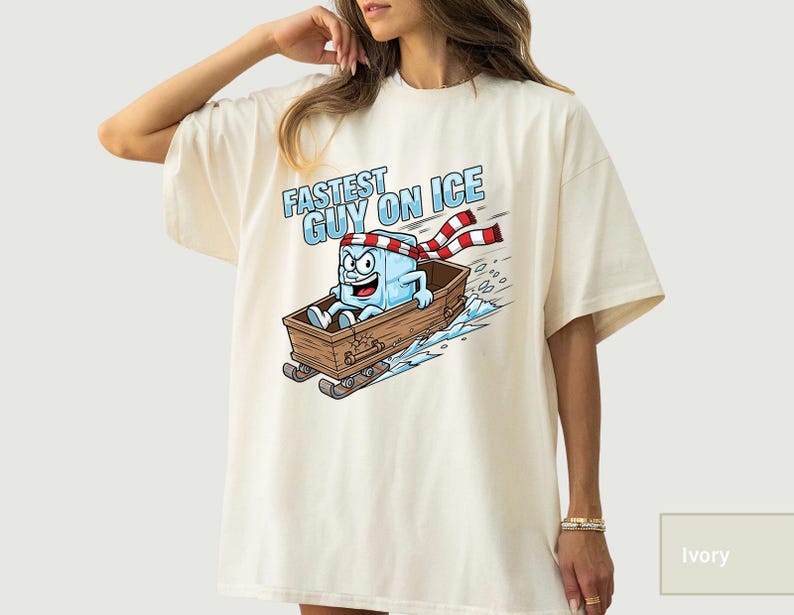 May include: Ivory t-shirt with a cartoon ice cube character in a wooden sled, with the text "FASTEST GUY ON ICE." The ice cube wears a red and white striped scarf and headband. The design includes blue ice and snow.