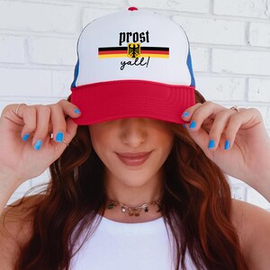 May include: A white, red, and blue trucker hat with the text "prost y'all!" and a German flag design. The hat has a red brim, a white front panel, and a blue mesh back. The text is in black script.