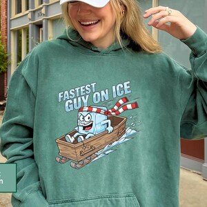 May include: Light green hoodie featuring a cartoon ice cube character in a wooden sled, with the words "Fastest Guy on Ice". The ice cube wears a red and white striped scarf and a headband.