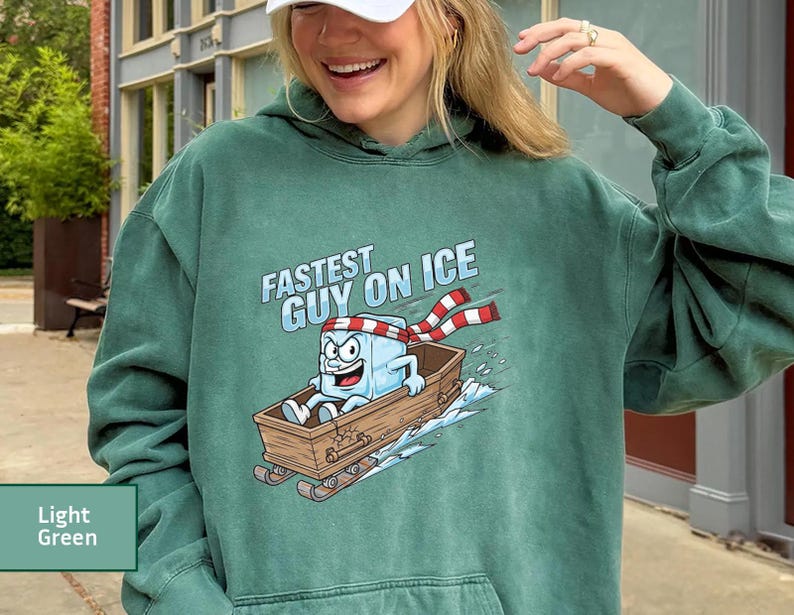 May include: Light green hoodie featuring a cartoon ice cube character in a wooden sled, with the words "Fastest Guy on Ice". The ice cube wears a red and white striped scarf and a headband.