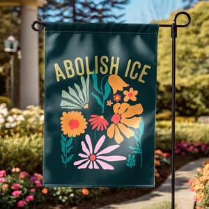 May include: A dark green garden flag with the words "ABOLISH ICE" in gold lettering. The flag features a colorful floral design with orange, pink, and yellow flowers and green leaves. The flag is displayed on a black metal garden stake.