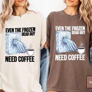 May include: Ivory and espresso-colored t-shirts with the text "EVEN THE FROZEN DEAD GUY NEED COFFEE". The graphic shows a hand emerging from an ice block reaching for a steaming coffee cup. The shirts are casual wear.