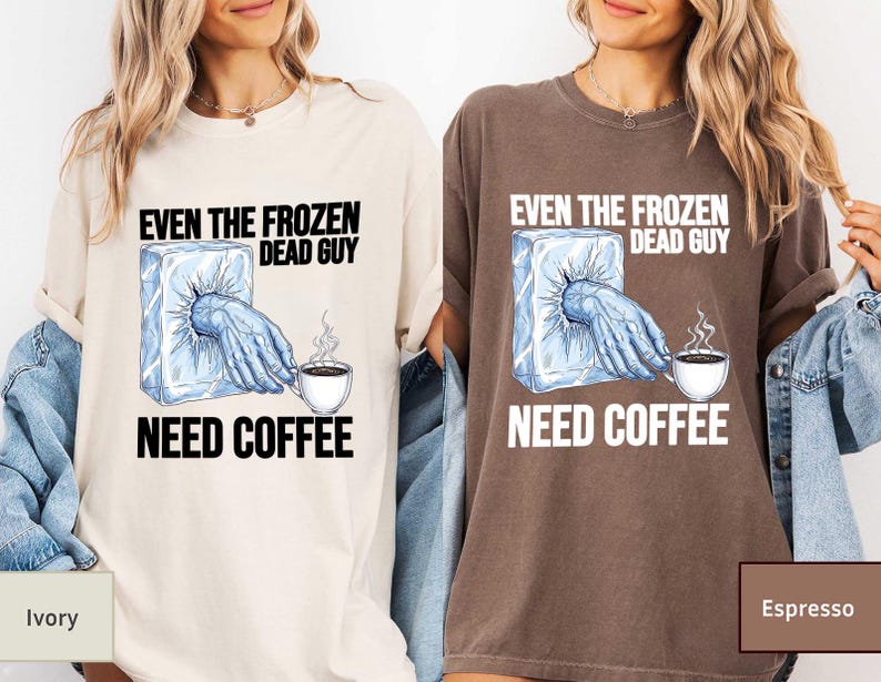 May include: Ivory and espresso-colored t-shirts with the text "EVEN THE FROZEN DEAD GUY NEED COFFEE". The graphic shows a hand emerging from an ice block reaching for a steaming coffee cup. The shirts are casual wear.