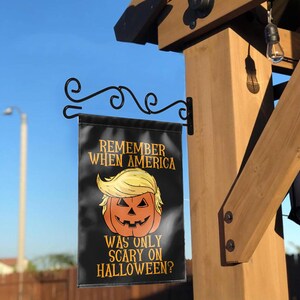 Puede incluir: Una bandera de jard&iacute;n negra de Halloween con el texto "REMEMBER WHEN AMERICA WAS ONLY SCARY ON HALLOWEEN?" presenta una calabaza con pelo rubio. La bandera est&aacute; colgada de un soporte de metal negro en una estructura de madera.