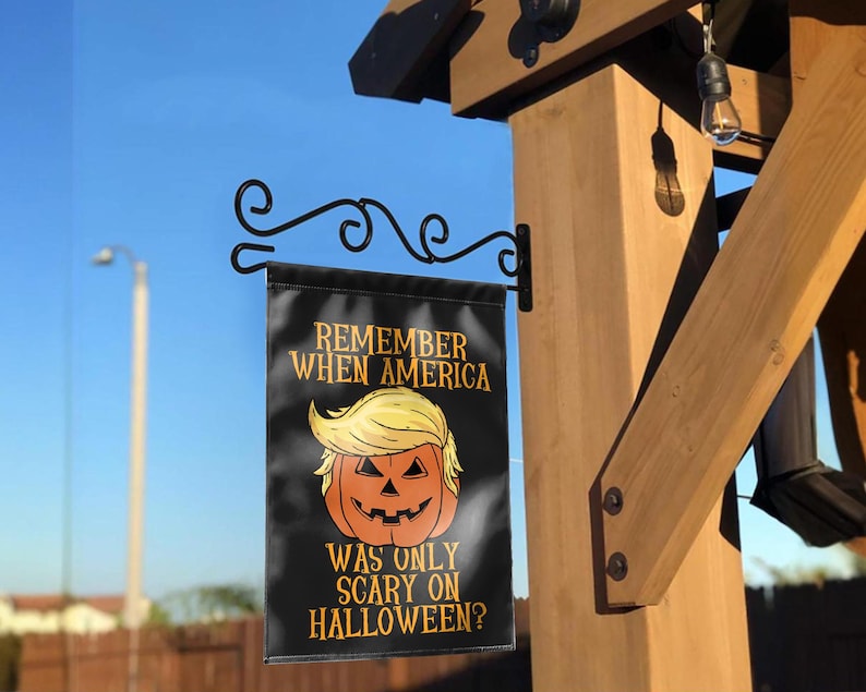 Puede incluir: Una bandera de jard&iacute;n negra de Halloween con el texto "REMEMBER WHEN AMERICA WAS ONLY SCARY ON HALLOWEEN?" presenta una calabaza con pelo rubio. La bandera est&aacute; colgada de un soporte de metal negro en una estructura de madera.