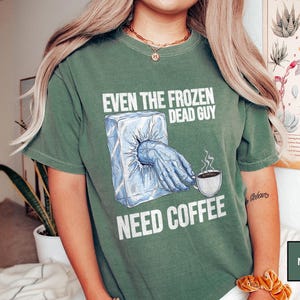 May include: Moss green t-shirt with the text "EVEN THE FROZEN DEAD GUY NEED COFFEE". The graphic shows a hand emerging from an ice block, reaching for a coffee cup. The shirt is a comfortable, casual style.