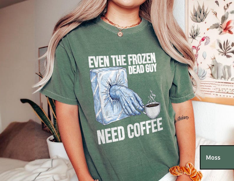 May include: Moss green t-shirt with the text "EVEN THE FROZEN DEAD GUY NEED COFFEE". The graphic shows a hand emerging from an ice block, reaching for a coffee cup. The shirt is a comfortable, casual style.