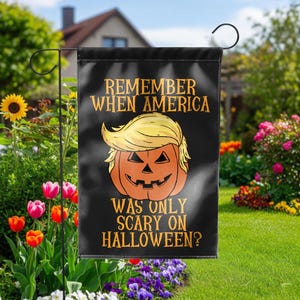 Puede incluir: Bandera de jard&iacute;n negra con un dise&ntilde;o de calabaza de Halloween con un peinado rubio. El texto dice: "REMEMBER WHEN AMERICA WAS ONLY SCARY ON HALLOWEEN?" La bandera se exhibe en un jard&iacute;n con flores coloridas y vegetaci&oacute;n.
