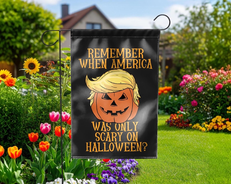Puede incluir: Bandera de jard&iacute;n negra con un dise&ntilde;o de calabaza de Halloween con un peinado rubio. El texto dice: "REMEMBER WHEN AMERICA WAS ONLY SCARY ON HALLOWEEN?" La bandera se exhibe en un jard&iacute;n con flores coloridas y vegetaci&oacute;n.