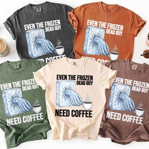 May include: A collection of five short-sleeved t-shirts in various colors, each featuring the text "EVEN THE FROZEN DEAD GUY NEED COFFEE." The graphic shows a hand emerging from ice, reaching for a coffee cup.
