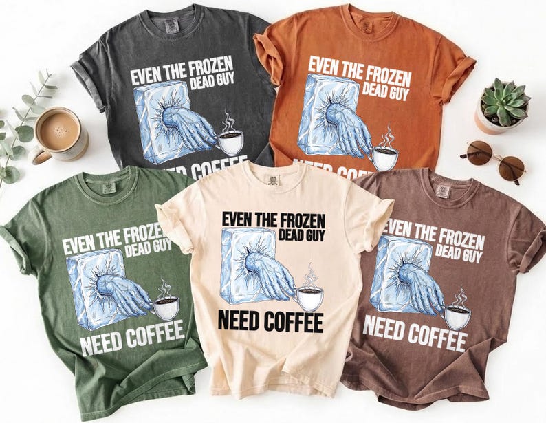 May include: A collection of five short-sleeved t-shirts in various colors, each featuring the text "EVEN THE FROZEN DEAD GUY NEED COFFEE." The graphic shows a hand emerging from ice, reaching for a coffee cup.