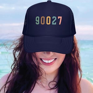 May include: Navy blue trucker hat with the number "90027" printed in retro colors. The hat has a mesh back and a curved brim. The background shows a beach scene with blue water and sky.