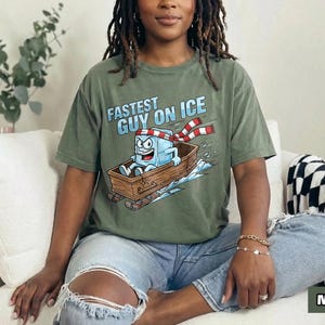 May include: An olive green t-shirt featuring the text "FASTEST GUY ON ICE" and a cartoon ice cube character on a sled. The ice cube wears a red and white scarf. The word "MOSS" is in a green rectangle.