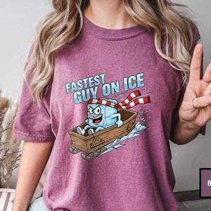 May include: Berry-coloured t-shirt with a cartoon ice cube character in a wooden sled, and the text "Fastest Guy On Ice." The ice cube wears a red and white striped scarf and has an excited expression. The design is printed on a berry-coloured shirt.