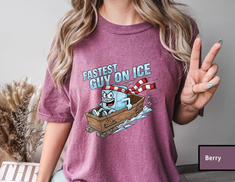 May include: Berry-coloured t-shirt with a cartoon ice cube character in a wooden sled, and the text "Fastest Guy On Ice." The ice cube wears a red and white striped scarf and has an excited expression. The design is printed on a berry-coloured shirt.