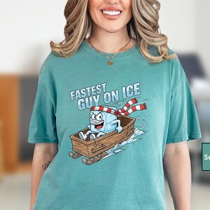 May include: Seafoam green t-shirt featuring a cartoon ice cube character in a wooden sled, wearing a red and white striped scarf. The text "FASTEST GUY ON ICE" is above the graphic.