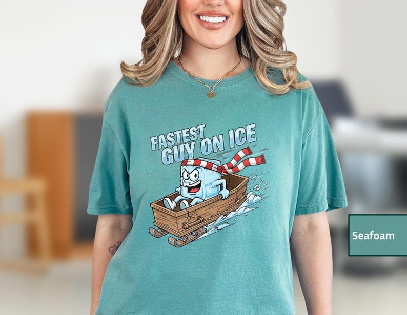 May include: Seafoam green t-shirt featuring a cartoon ice cube character in a wooden sled, wearing a red and white striped scarf. The text "FASTEST GUY ON ICE" is above the graphic.
